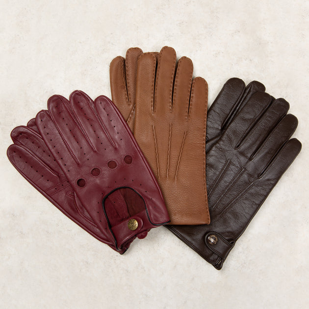 Three leather gloves arranged overlapping on a light beige textured background: one burgundy glove with a cuff opening and snap, one tan glove, and one dark brown glove.