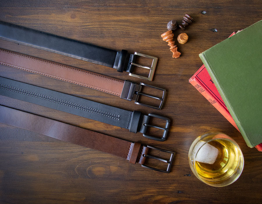 Four leather belts with buckles on a wooden surface, by a glass of whiskey and books.