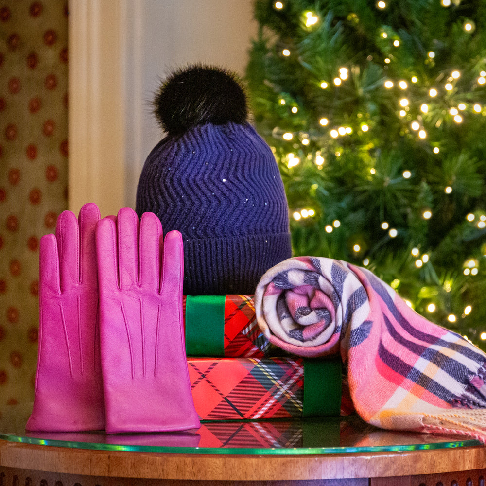 Women's pink leather gloves, a pink plaid scarf and navy blue bobble hat with wrapped gift presents in a cosy setting with Christmas tree