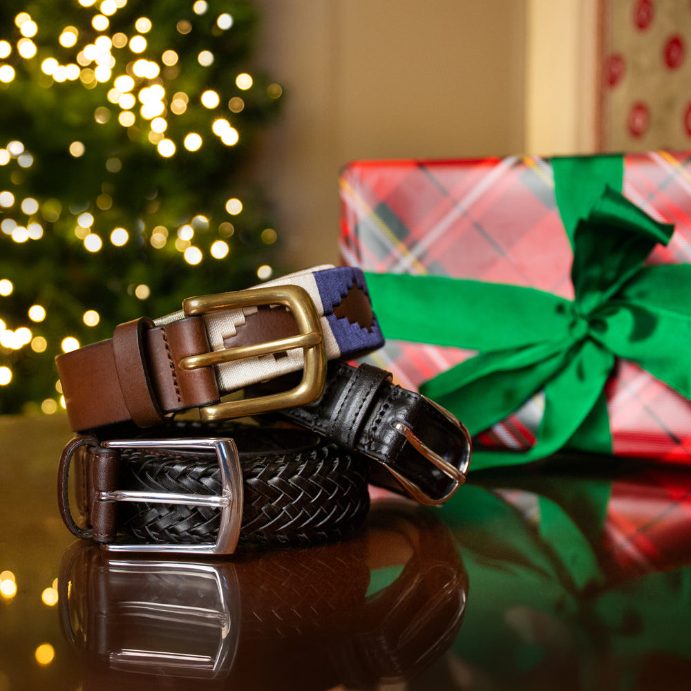 Three men's leather belts on a table with a blurred Christmas tree and a wrapped gift box in the background.