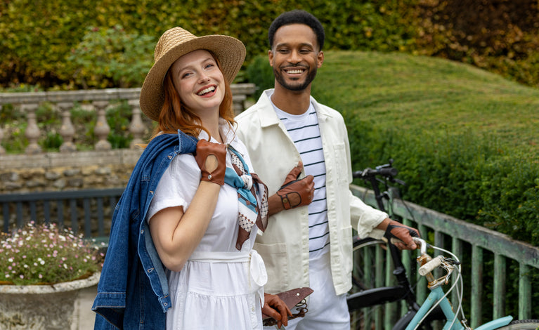 A smiling couple posing outdoors in a garden setting. Wearing leather gloves in brown shades. 