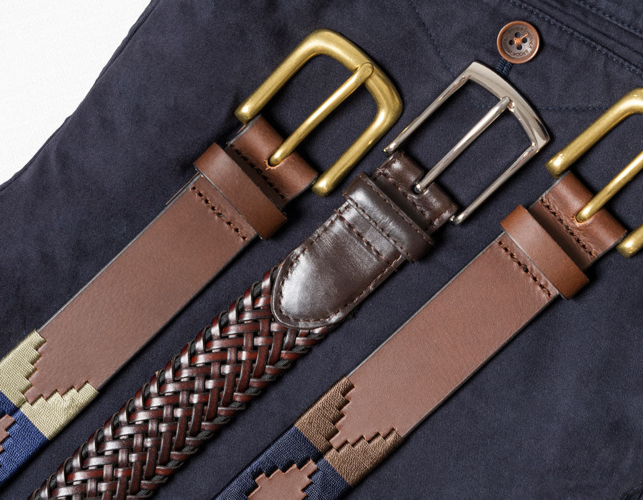 Three brown leather belts with different buckle finishes and woven details, laid out diagonally on a dark navy blue fabric background.
