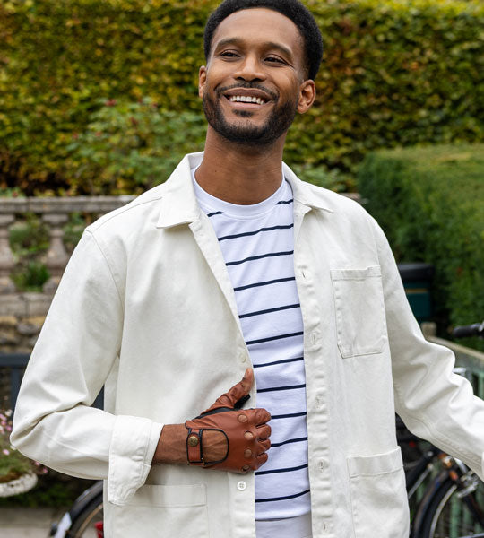 Man wearing a fingerless leather driving gloves in cognac, standing outdoors with greenery in the background
