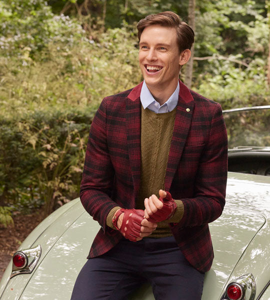 Man wearing leather fingerless driving gloves in berry with check jacket outside leaning on a boot of a classic car
