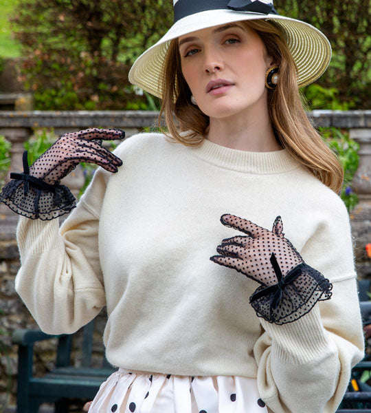 Woman wearing a cream sweater with black lace gloves and a wide-brimmed hat outdoors.