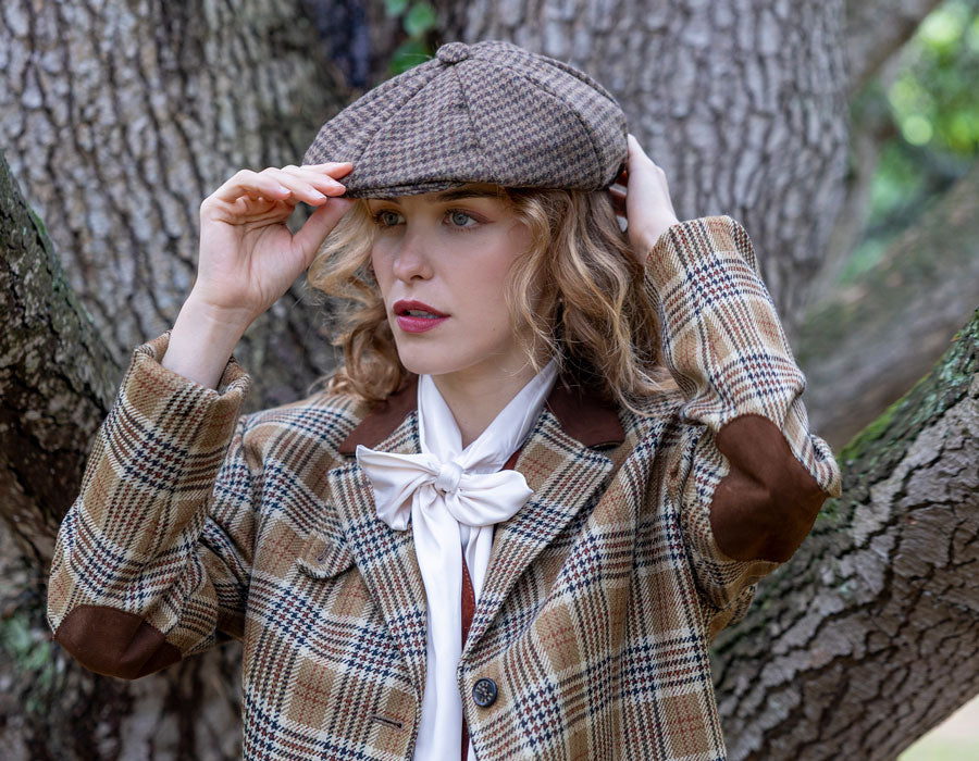 A woman wearing a brown and beige plaid flat cap, outdoors in front of a large tree trunk.