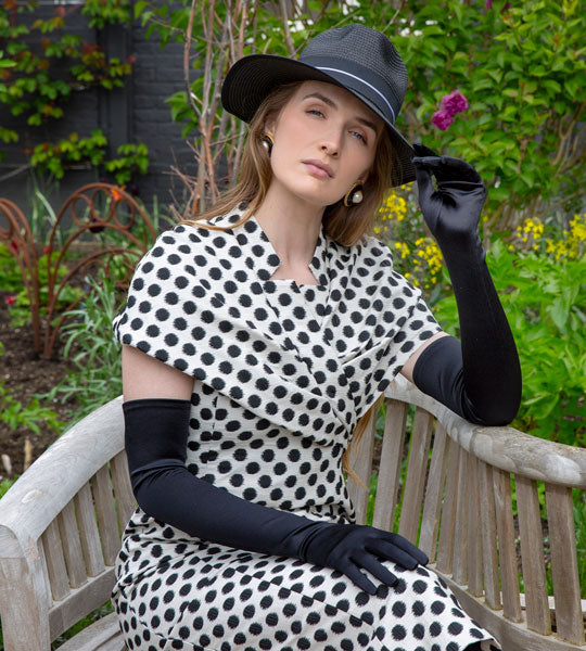 Woman in a polka dot dress and black long satin gloves sitting on a bench in a garden.