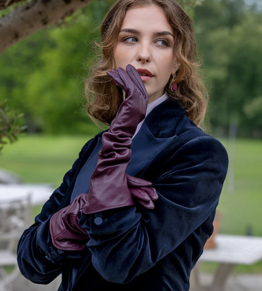 A woman outdoors wearing long burgundy leather gloves and a dark navy velvet jacket, with a blurred green park background.