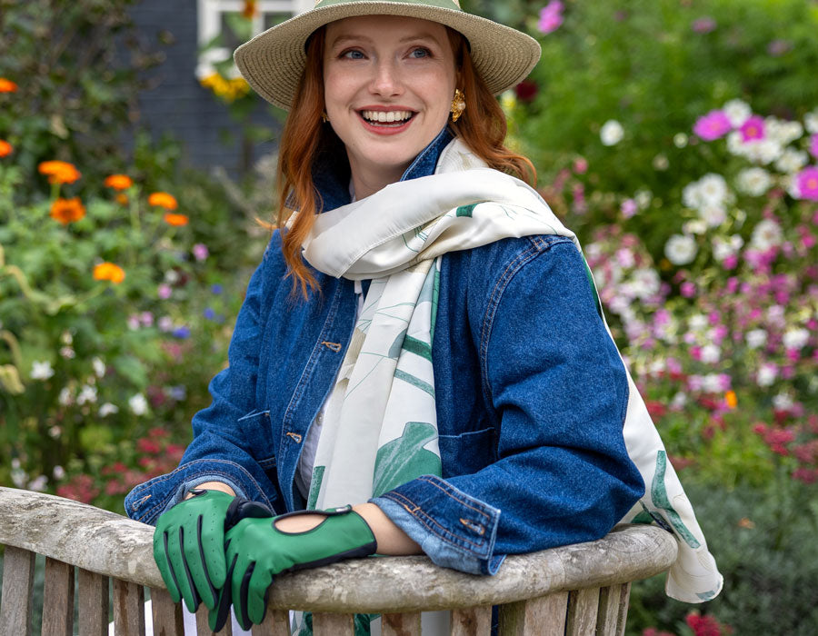 Woman in a garden wearing a hat, gloves, and a scarf with flowers in the background