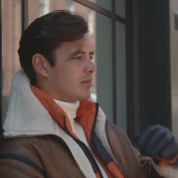Man wearing cashmere-lined leather gloves in navy/tangerine with a fleece-lined coat in brown outside an apartment building